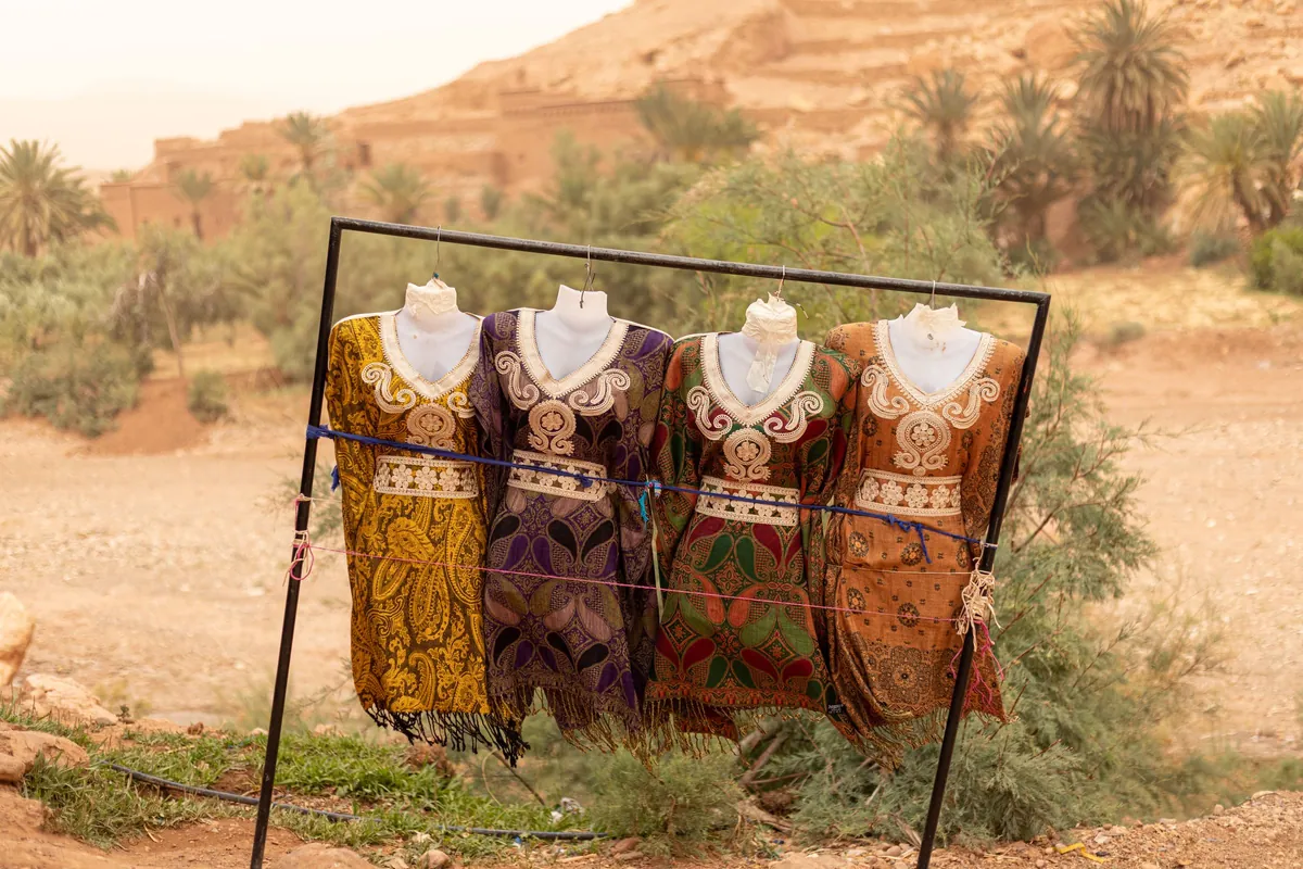 Quatre caftans brodés ocre, rouge et violet sur des bustes de mannequin, portant installé en plein air près de palmiers