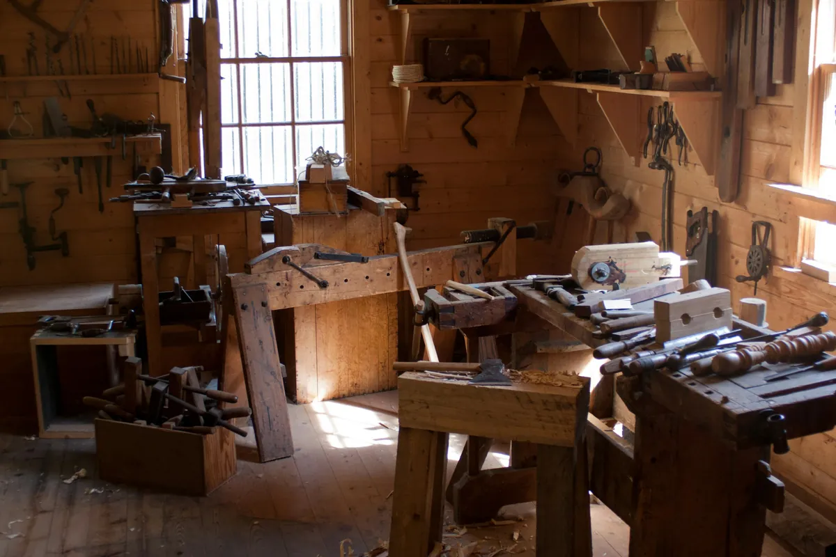 Atelier de menuiserie avec établis en bois, outils accrochés au mur et rabots, éclairé par les fenêtres
