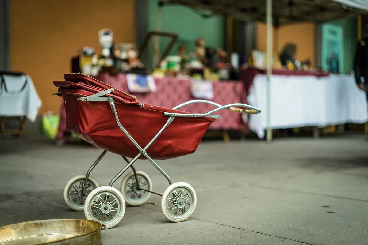 Ancien landau de poupée rouge à roues blanches posé au sol devant les stands d’un marché aux puces