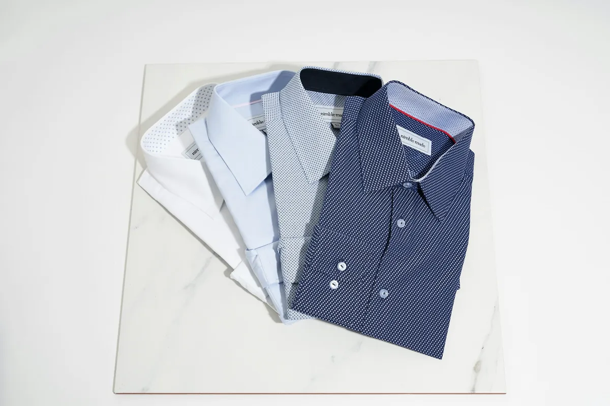 Quatre chemises pour homme pliées en éventail sur du marbre blanc : blanche, bleu clair, rayée et bleu marine