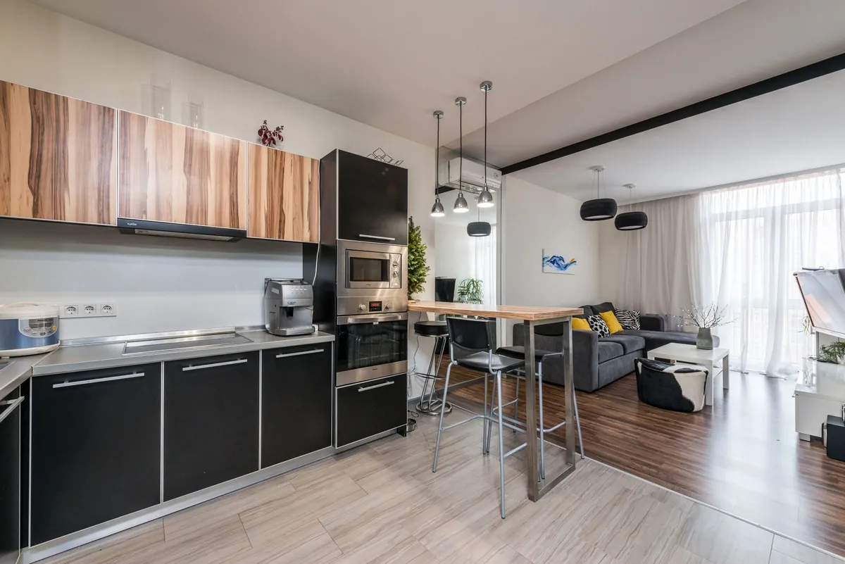 Appartement à pièce de vie ouverte : cuisine noire et bois, îlot avec tabourets hauts, grandes fenêtres