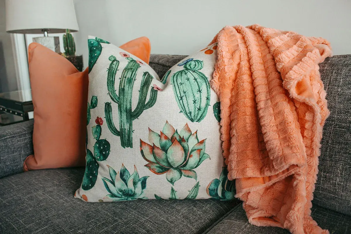 Coussin imprimé de cactus, coussin orange uni et plaid orange à franges sur un canapé gris