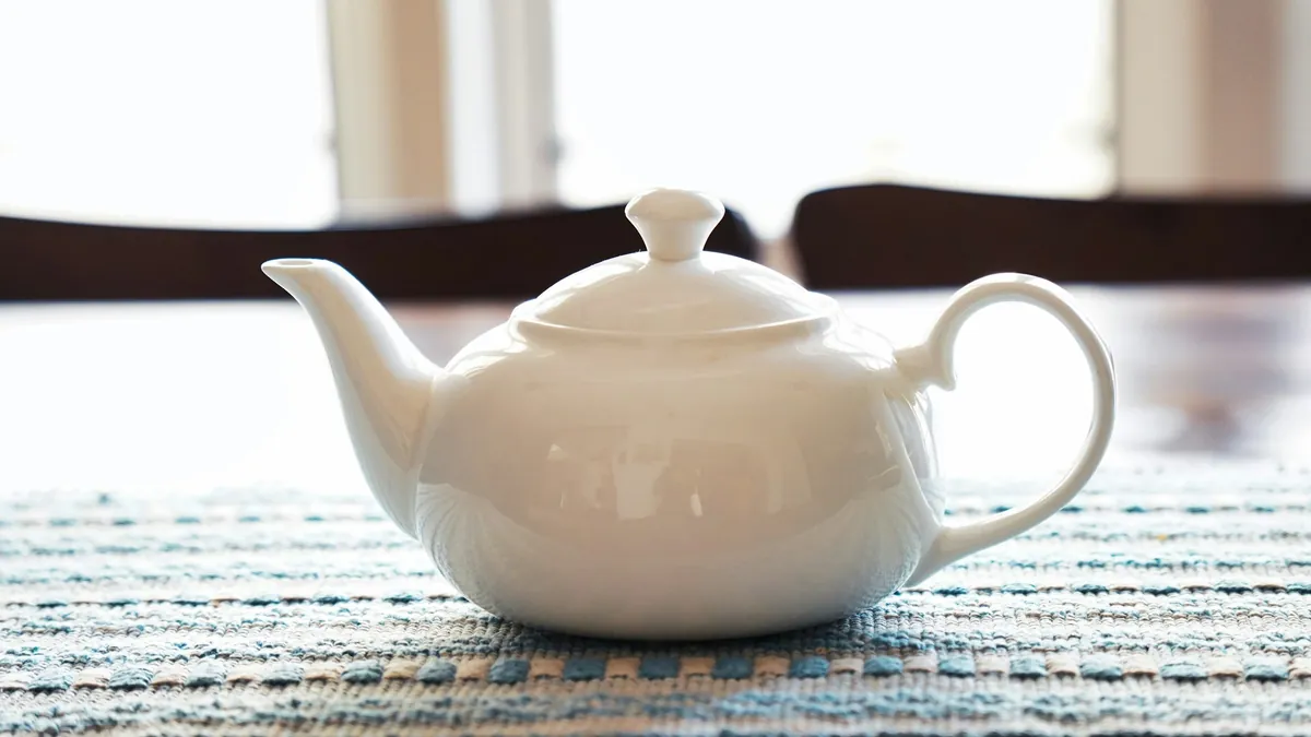 Théière en porcelaine blanche sur un set de table tissé bleu et blanc, devant une fenêtre lumineuse