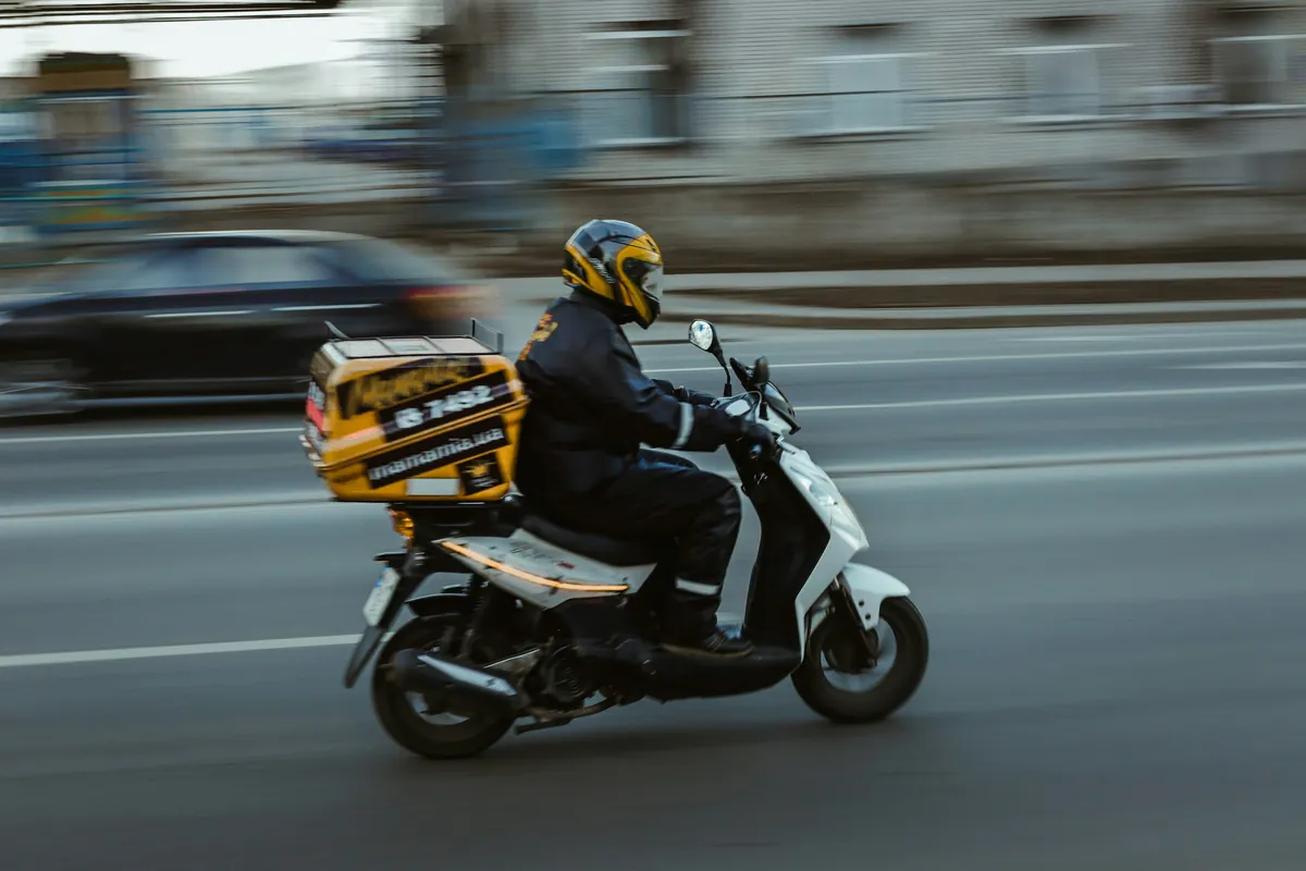 Livreur casqué sur un scooter équipé d’un coffre de livraison jaune, roulant dans une rue