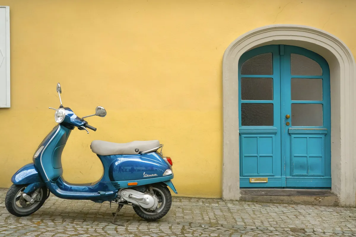 Scooter bleu de style rétro garé sur des pavés devant une façade jaune et une porte cintrée bleue
